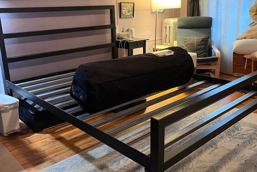 a bedroom with a black slatted bed frame and a rolled tempur cloud mattress in a black bag sitting on top during the setup process