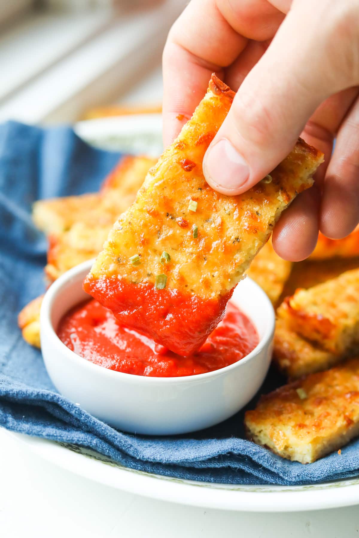 A hand holding a piece of cheese bread that has marinara sauce on the end of it. There is a white cup filled marinara sauce below the piece of bread.